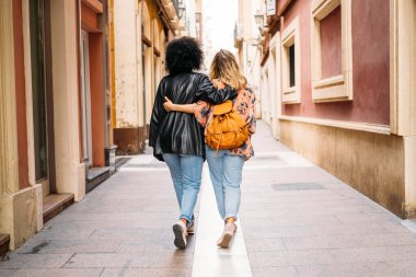 Dar bir caddede kol kola yürüyen iki farklı kadın arkadaş.