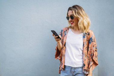 Genç sarışın kadın gülümsüyor, güneş gözlüğü takıyor, cep telefonundan içeriğini kontrol ediyor.