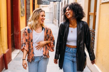 Dar bir sokakta birlikte yürüyen, mutlu bir anı paylaşan iki farklı kadın.