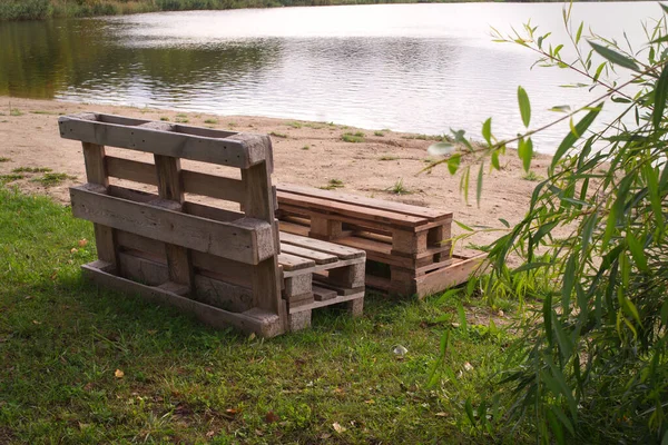 Outdoor furniture made from wood pallets on the shore of a tranquil a lake surrounded by trees and greenery in summer sunshine