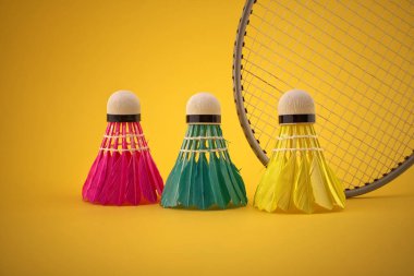 Badminton tüylü mekikleri yeşil, sarı ve pembe renkte badminton raketinin yanında fotokopi alanı olan sarı arkaplan üzerinde.