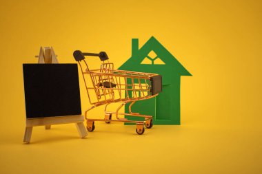 Ev ile mülkiyet kavramı, sarı arkaplan üzerinde küçük karatahta ve alışveriş arabası ve metin için ücretsiz kopya alanı