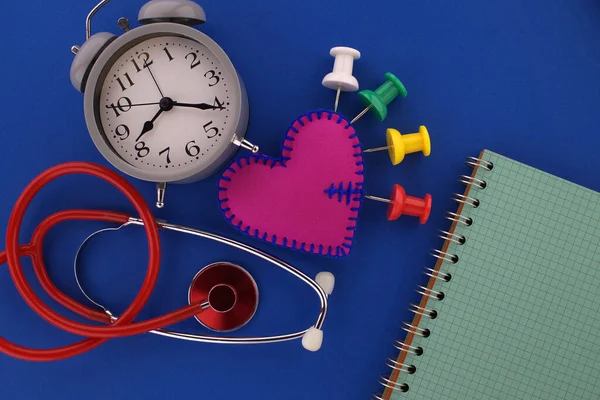 Heart pain, medical concept with multi color push pins stuck into a felt heart symbolising pain intensity, stethoscope with clock and open wire bound notebook for your text