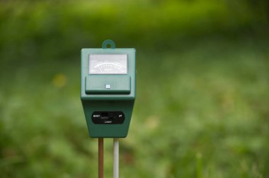 Çimenli bir açık hava bahçesinde toprak nem ve pH değerlerini ölçmeye hazır küçük yeşil bir el bilgisayarı duruyor. Hızlı ve basit okuma gerektiren bahçıvanlık, tarım ve peyzaj projeleri için ideal.