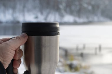 Bir el paslanmaz çelik bir seyahat kupasını siyah bir kapakla kavrıyor, buzlu, sakin, karlı bir zemine dayanıyor. Soğuk bir açık hava sahnesinde samimi, taşınabilir sıcaklık..