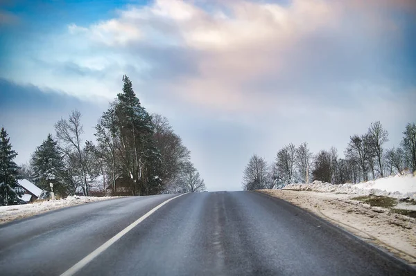 Sakin bir kış yolu, bulutlu mavi bir gökyüzünün altında karla kaplı ağaçlarla çevrili olan mesafeye uzanıyor. Islak asfalt, soğuk havada sükuneti ve seyahati davet eden son buzun eridiğini ima ediyor..