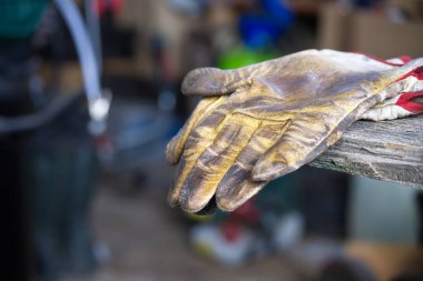 Çamurlu bir deri eldiven, hareketli bir atölyede, pürüzlü ve yıpranmış bir bankın üzerine örtünür. Yıpranmış dokusu ve kiri ağır işçiliği ve usta zanaatkarlığı taşıyor..
