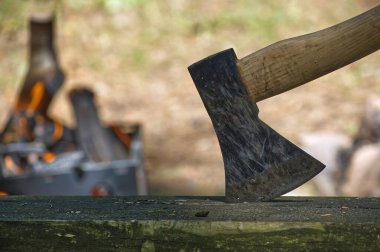 Yıpranmış balta kafası parlayan bir kamp ateşinin yanında tahta bir kütüğe çarpmış, kırsal bir doğa yaşamını çağrıştırıyor, odun doğrama, kamp sırasında sıcaklık ve el işçiliği, çiftlik işleri, ya da sonbahar akşamları