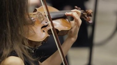 Kadın kemancının, klasik tiyatro sahnesinde konser sırasında arka planda senfoni orkestrasının sahne aldığı kemanlar çalarken yakın plan çekimleri yapılıyor..