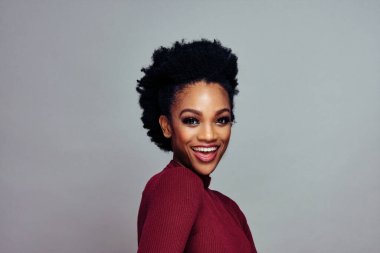 Kıvırcık saçlı, gri arka planda kırmızı kazak giyen mutlu gülümseyen genç bir kadının portresi.