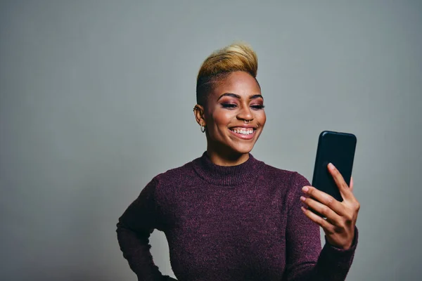 Smiling young female hipster looking away while using smart phone against gray background