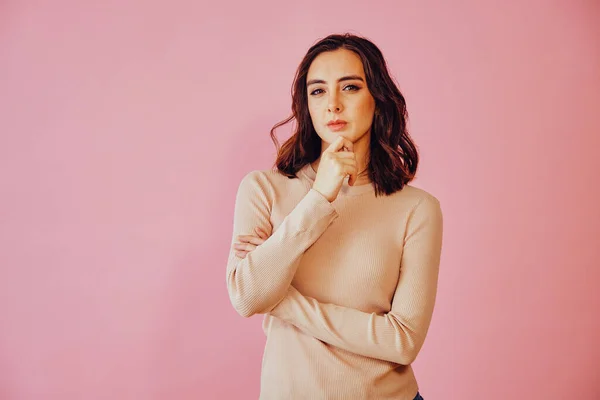 Eli çenesinde ve kolları bağlı düşünceli bir kadın pembe arka grubuna karşı kameraya bakıyor.