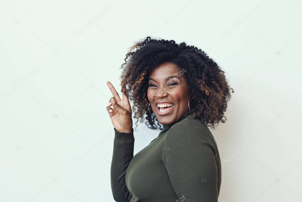 Side view of cheerful curly haired woman showing peace sign against white background