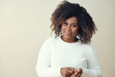 Kıvırcık saçlı, kahverengi röfleli gülümseyen Afro kadın beyaz arka planda kameraya bakıyor.