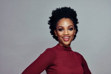 Kıvırcık saçlı, gri arka planda kırmızı kazak giyen mutlu gülümseyen genç bir kadının portresi.