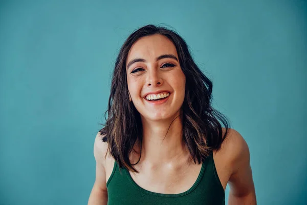 Güzel, cana yakın bir kadın kameraya bakıp gülüyor mavi arka planda kolsuz bluz giyiyor.