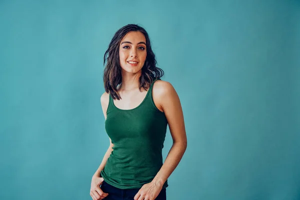Kolsuz tişörtlü, elleri ceplerinde, gülümseyen ve kameraya bakan genç bir kadın.