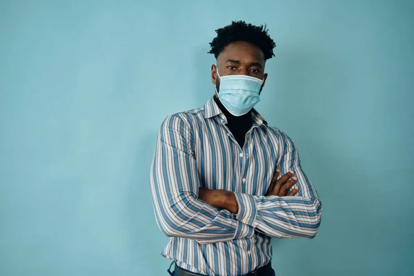 Mavi arka planda izole edilmiş kolları bağlı tıbbi maske takan Afro-Amerikan bir adam kameraya bakıyor.