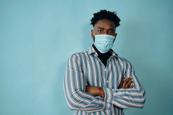 african american man in medical mask with arms folded standing isolated over blue background, looking at camera
