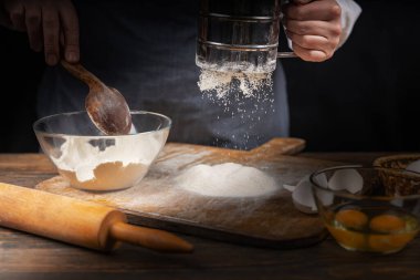 Women's hands, flour and dough. A woman, in an apron prepares dough for homemade baking, a rustic home cozy atmosphere, a dark background with unusual lighting.