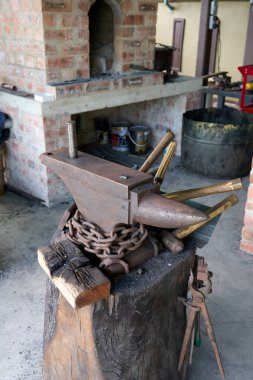 Sağlam, kırsal bir örs, ahşap tezgahında duran çeşitli çekiç ve aletlerle çevrili geleneksel bir demirci atölyesinde ortaya çıkıyor. Örsün etrafına ağır bir zincir örülmüş. Otantik çalışma atmosferine ekleniyor.. 