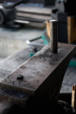 Yakın plan, açılı bir bakış açısı, bir demircinin örsünün yıpranmış yüzeyine odaklanarak, dokusu ve kullanılmış durumunu gösterir. Küçük, yumruklanmış metal bir parça örsün yüzünde duruyor. Pritchel deliğine bir yuvarlak yumruk aleti yerleştiriliyor.