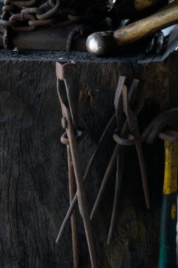 Bu yakın çekim, ahşap örs tezgahının yanında asılı duran iyi kullanılmış, rustik demirci maşası dizisini yakalıyor. Karanlık.,