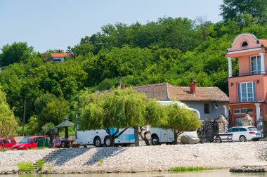 Güneşli bir günde nehir kıyısındaki ormanın yakınındaki arabaların ve binaların manzarası.