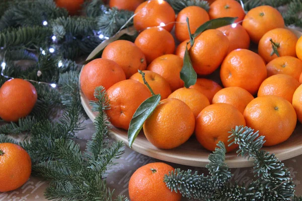 Small tangerines close-up on a plate decorated with coniferous green branches and a garland — Stock Photo, Image