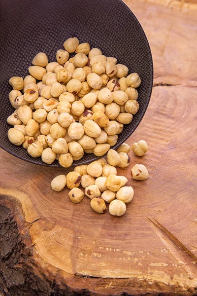 The kernels of the peeled, shell-less hazelnuts fall from a dark cup on a wooden background. — Foto Stock