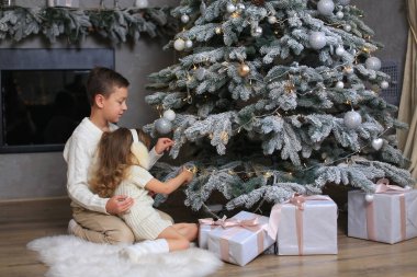 Çocuklar - beyaz örgü süveter giyen bir erkek kardeş ve beyaz örgü elbise ve beyaz kürk kulaklıklı bir kız kardeş Yeni Yıl ağacını süslüyor.