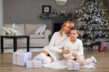 Beyaz örülü bir elbise giyen anne, beyaz kazaklı oğluyla birlikte suni beyaz kürkün üzerindeki bir odada oturuyor. Kutuların arkasında hediyeler ve yeni yıl ağacı var. Mutlu Noeller..