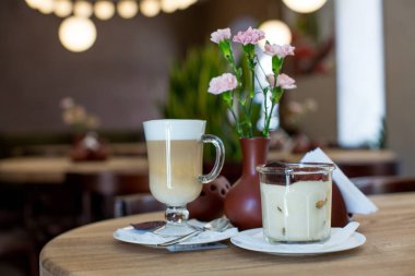 Tiramisu in a transparent glass, lat, pieces of dark chocolate on a wooden table against the background of a ceramic vase with pale pink carnations and a holder with napkins