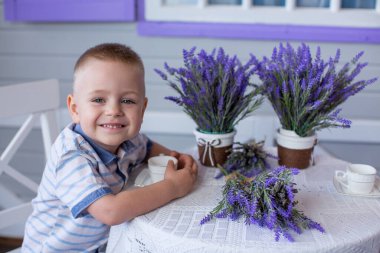 Provence 'ın manzarasında bir çocuk masada oturuyor. Ön cephenin arka planında kepenkli lavanta buketleri olan beyaz masa ve sandalye. Masanın üzerinde beyaz bir fincan ve çay tabağı var.