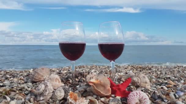 海と雲の青空を背景にシェルの海岸に赤いドライワインスタンドで満たされた2つのメガネ — ストック動画