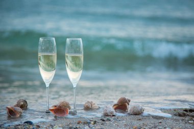 Kum, deniz kabuğu ve dalgaların arka planında beyaz köpüklü şaraplı iki bardak.