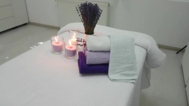 Gevşek balmumlu üç mum, sarılmış lavanta renkli havlular ve masaj salonundaki kanepede çiçek açan bir demet lavanta.