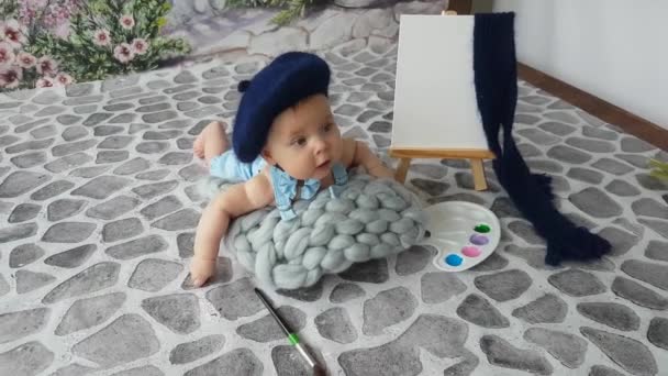 青いベレー帽をかぶった5ヶ月のアーティストの少年がパレットとイーゼルで舗装に横たわっています 彼は本当に描きたいが まだできない — ストック動画