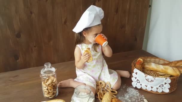 一个身穿白色衣服头戴烘焙帽的三岁小女孩吃了一个大百吉饼 并在烘焙的背景下喝着果汁 — 图库视频影像