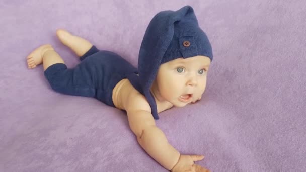 一个穿着蓝色侏儒服装的五个月大的男孩躺在紫色的床罩上 — 图库视频影像