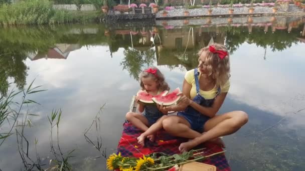 妈妈和四岁的金发碧眼的女儿在河边的一个钓鱼平台上吃西瓜 — 图库视频影像