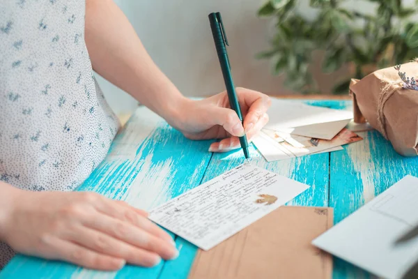 A woman writes a letter on a postcard. Hands holding a pen close-up. Blue wooden table with a parcel in the background. The concept of postcrossing.