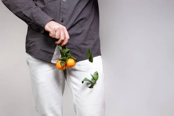 Concept of male sexual diseases. A man at the level of the genitals holds tangerines or oranges on a twig, from which the leaves fall off. Testicular cancer. — Stock Image Concept Male Sexual Diseases Man Level Genitals Holds Tangerines Oranges Stock Picture