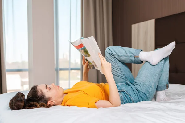 Reading and education. A woman reads a magazine lying on the bed. Indoor. Side view.