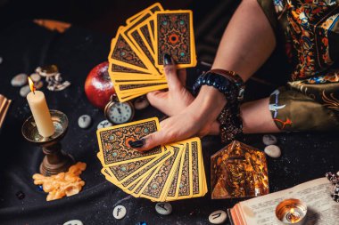 Astroloji ve ezoterizm. Bir falcının elinde Tarot kartları hayranı var. Masanın üzerinde rünler ve sihirli cam piramit var. Elleri yakın çekim. Üst görünüm.