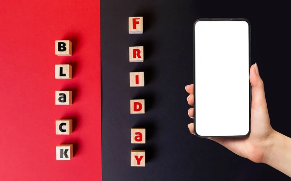 Black friday concept. On a black and red background are wooden cubes with the inscription black Friday. Flat lay. A woman\'s hand is holding a smartphone. Mock up.