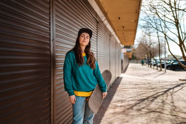 Genç hippi bir kadın elinde kaykayla poz veriyor. Arka planda kapalı dükkanları olan bir cadde var. Uzayı kopyala Spor yaşam tarzı ve sokak kültürü kavramı.