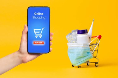 İlaç ve termometreyle dolu tıbbi maskeli bir alışveriş arabası. Bir kadının eli akıllı telefonu tutar. Sarı arka plan. Virüs koruması ve çevrimiçi ilaç alımı