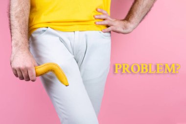 Impotence. A man in white jeans is holding a broken banana near his genitals with his legs together and his hand on his waist. Pink background. Close up. clipart