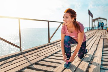 Spor. Spor giyimli mutlu yetişkin kadın, spor ayakkabılarının bağcıklarını bağlıyor. Arka planda iskele ve okyanus.
